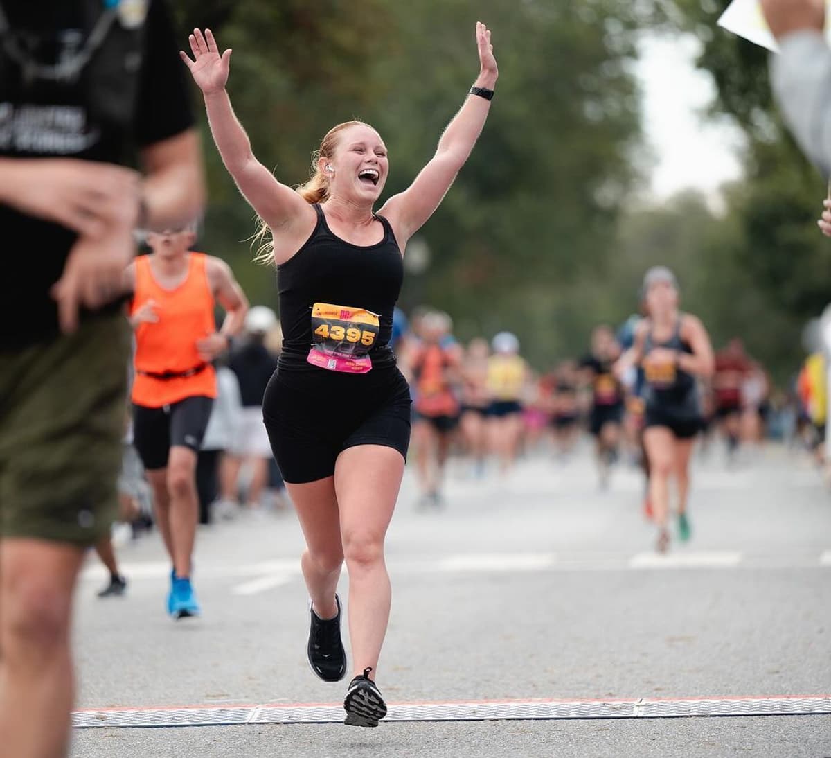 Runner in black outfit crosses the finish line with arms raised in celebration; other runners are visible in the background.