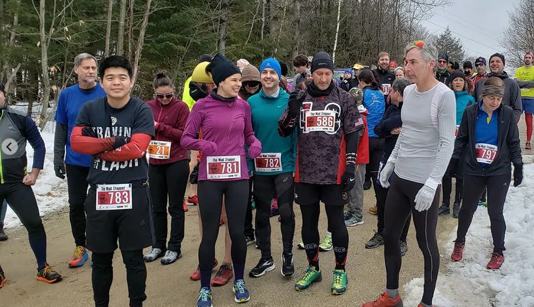 A group of runners wearing winter gear stand at a starting line on a snowy dirt path. They have numbered bibs and appear to be preparing for a race. Trees and more participants are in the background.