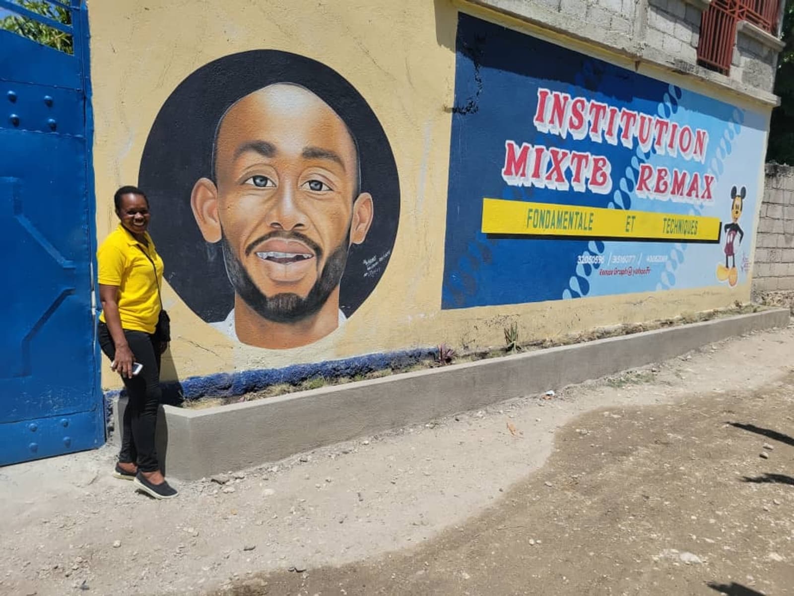 A person in a yellow shirt stands beside a mural featuring a portrait on a wall. The mural also includes text reading "Institution Mixte RBMAX," along with the words "Fondamentale et Secondaire." The scene is outdoors with a blue gate nearby.
