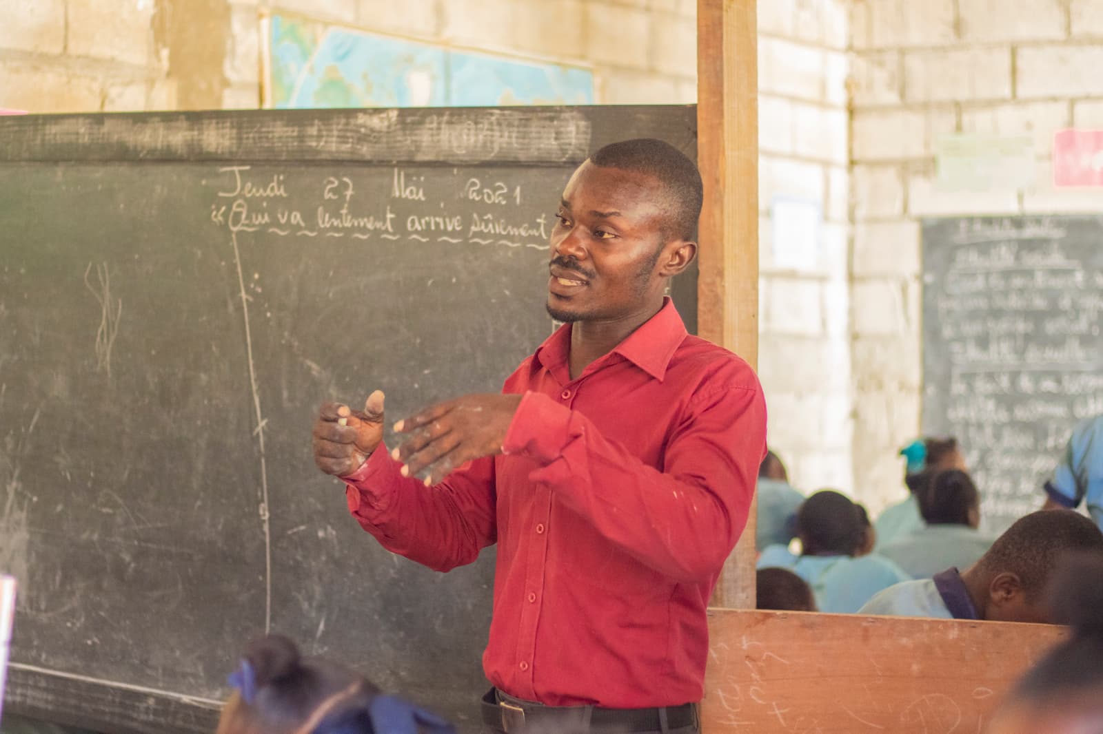 A teacher in a red shirt gestures while standing in front of a blackboard in a classroom. Students in blue uniforms are seated, listening attentively. The classroom has a rustic appearance with visible block walls.