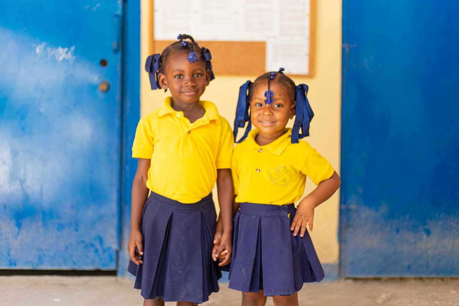 Two young girls in matching yellow shirts and navy skirts stand indoors. They both have hair styled with blue ribbons and are smiling at the camera, standing in front of a blue door and a bulletin board.