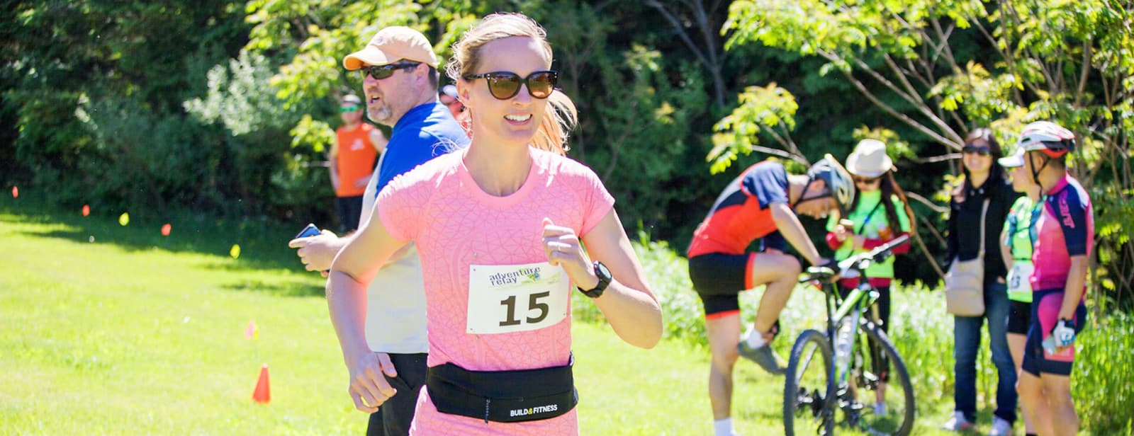 Woman wearing sunglasses and a pink shirt runs in a race with a number 15 bib. A group of people, some on bikes, stand in the background on a sunny day with greenery around.