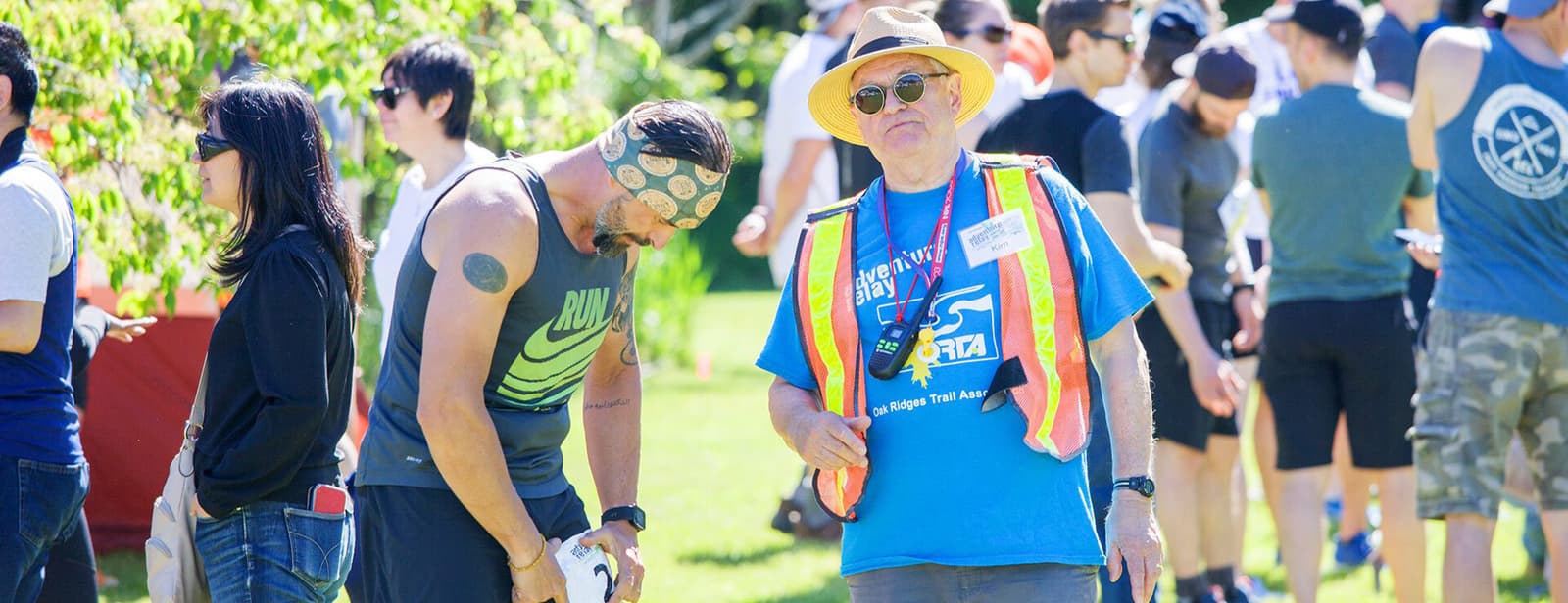A man in athletic gear adjusts his equipment while a man in a blue shirt and straw hat, wearing a high-visibility vest, stands beside him. A group of people is gathered in the background on a sunny day.