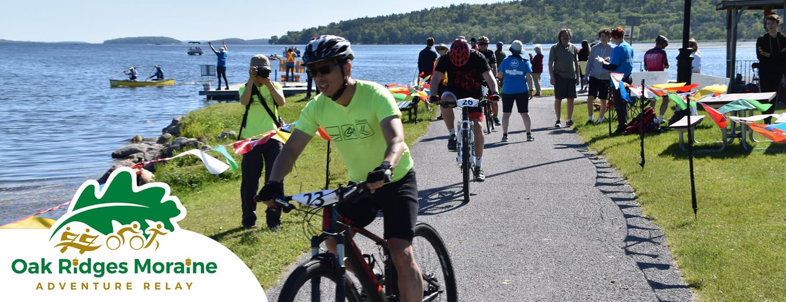 Cyclists participating in the Oak Ridges Moraine Adventure Relay ride along a lakeside path. The sunny scene features colorful flags, support crew, and a kayak on the water. Trees and hills are visible in the background.