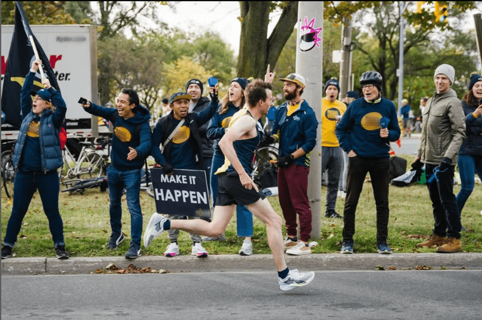 A runner in athletic gear sprints past a lively group of supporters, some cheering and holding a "MAKE IT HAPPEN" sign. The group stands on a sidewalk with trees and bikes in the background.