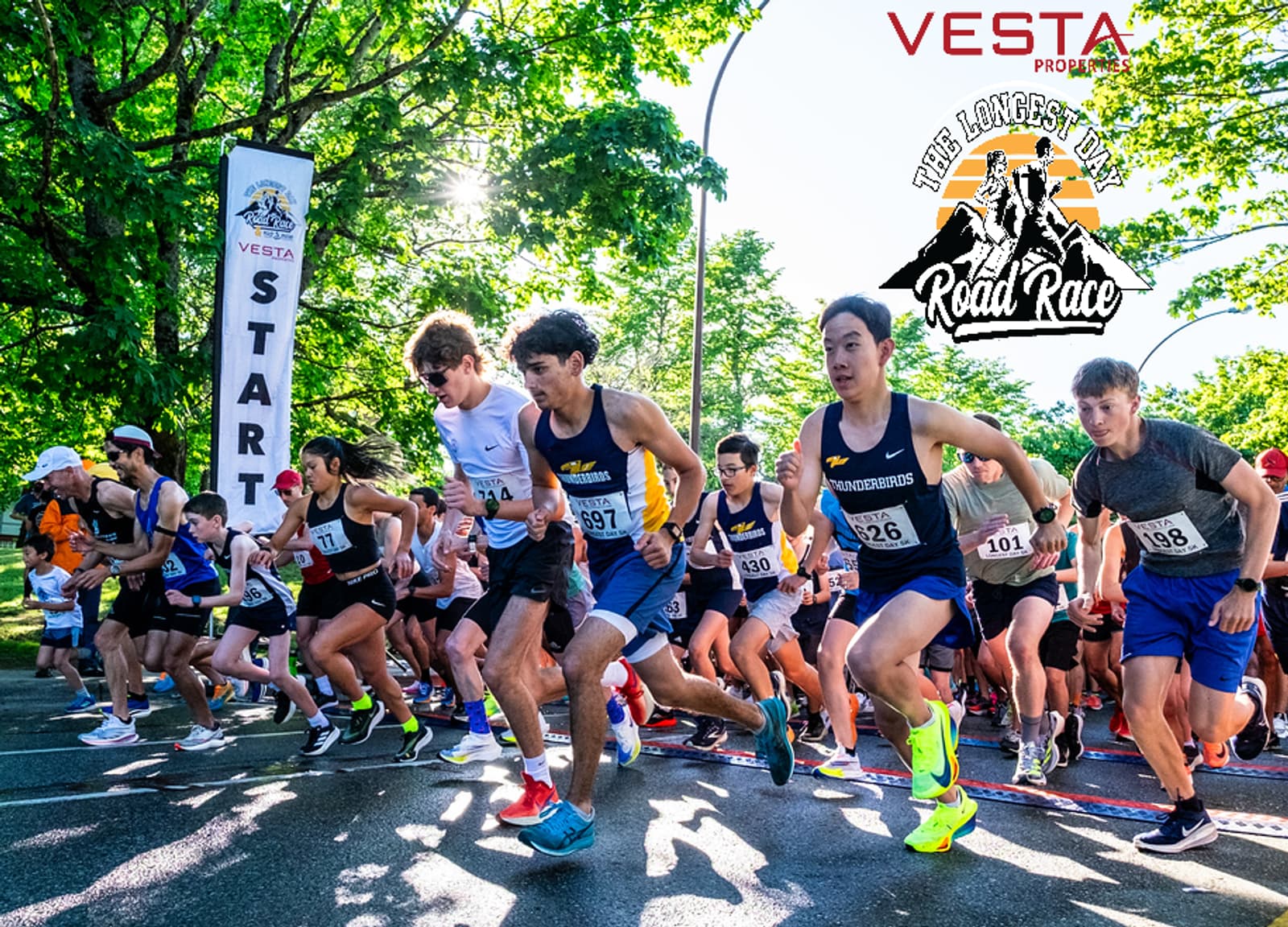 A group of runners in athletic gear launch from the starting line at an outdoor road race event, surrounded by trees. A "START" banner and "The Langley Road Race" logo are visible. Spectators line the background.