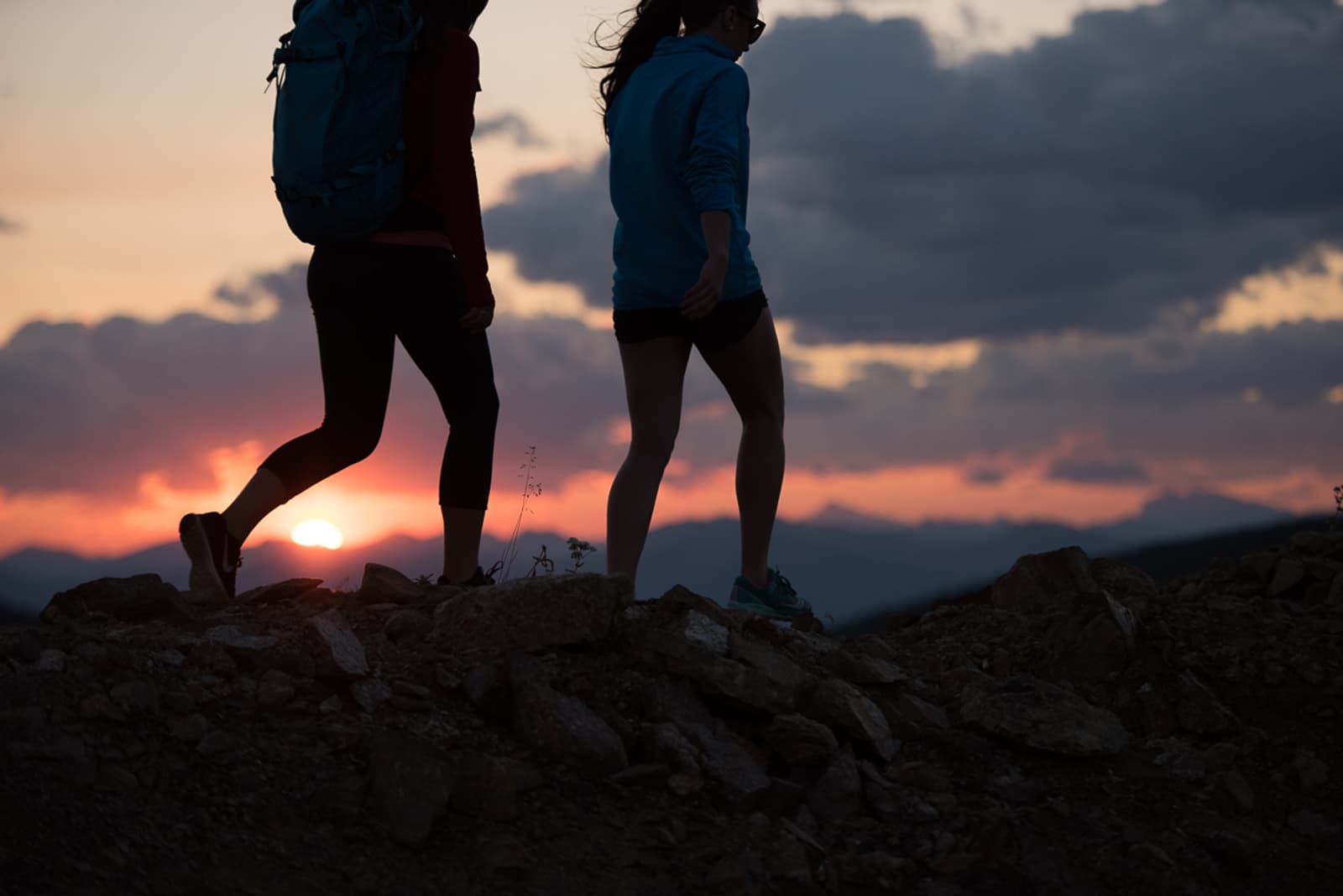 Two people hike along a rocky path at sunset, silhouetted against a colorful sky with clouds and distant mountains in the background.